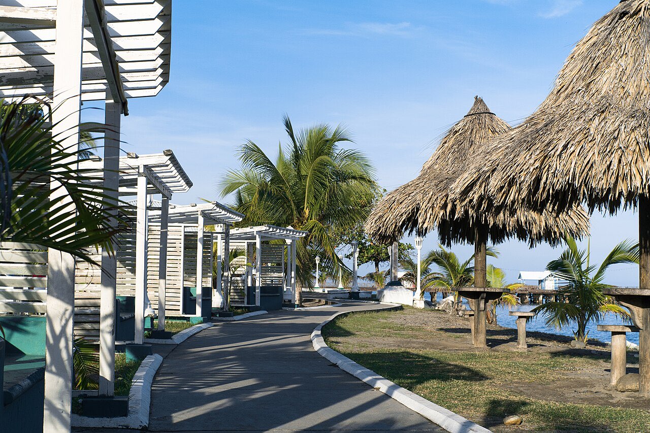 A paved waterfront walkway at La Ceiba lined with palms, thatched shelters and white pavilions